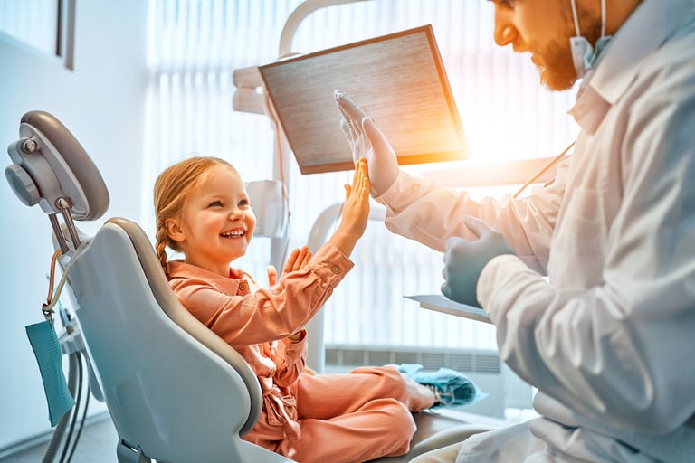 A little girl is sitting in a dentist's chair, giving a high five to the doctor and laughing. Children's dentistry. Sunlight.
