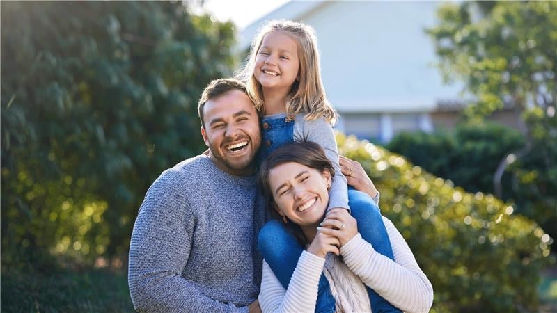 Happy family of four smiling outdoors with green trees and house in background